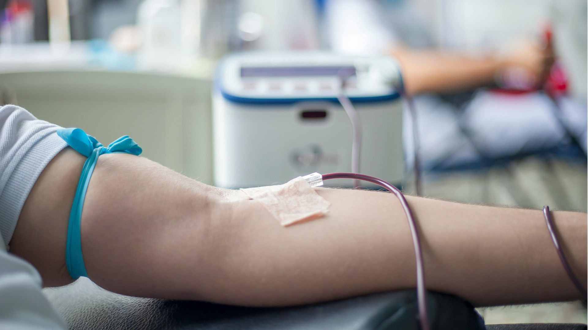 Arm receiving a blood donation through a needle and tubing, with a taped insertion site.