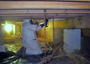 Person in a white protective suit working in a crawl space. Under beams, they use a tool near a chain.