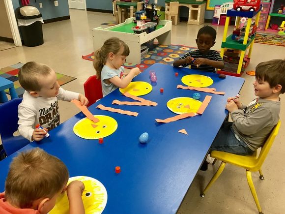 Children painting at a blue table with yellow paper plates in a classroom.