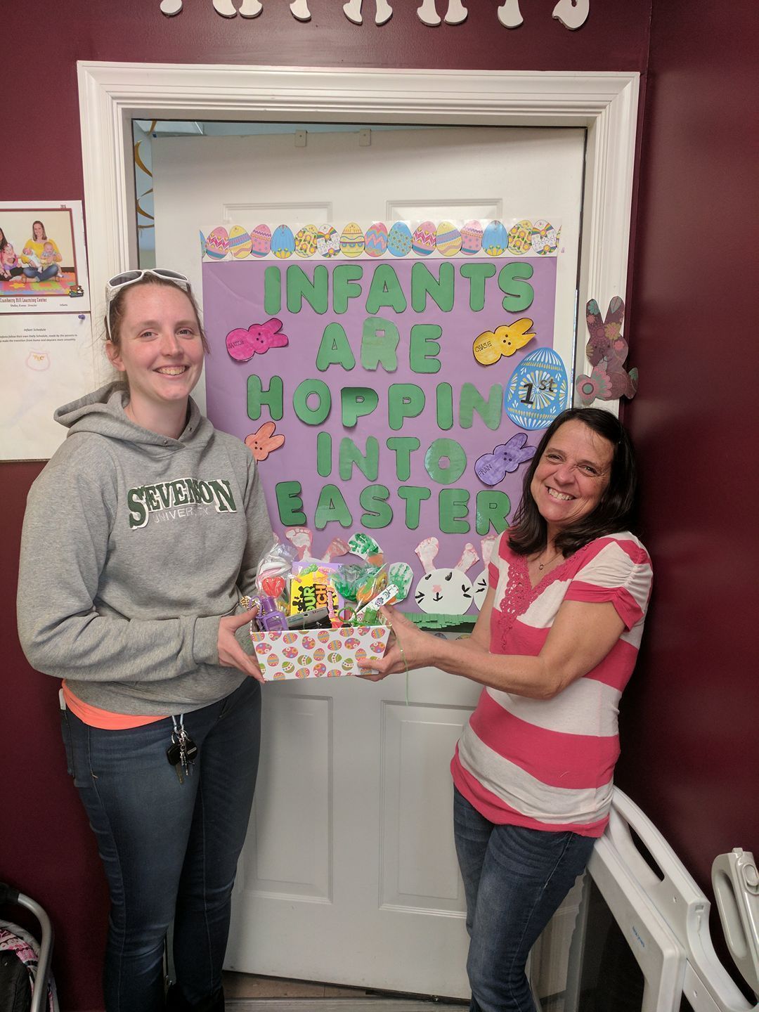 Two women hold a colorful sign reading “Infants are hopping into Easter” in a hallway.