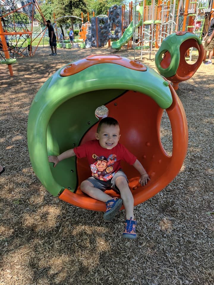 Child sitting inside a green and orange playground tube tunnel in a park, with swings and slides in the background.