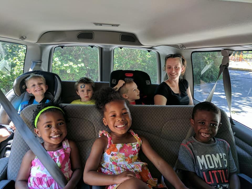 Six children seated in a car, smiling and buckled into their seats on a sunny day.