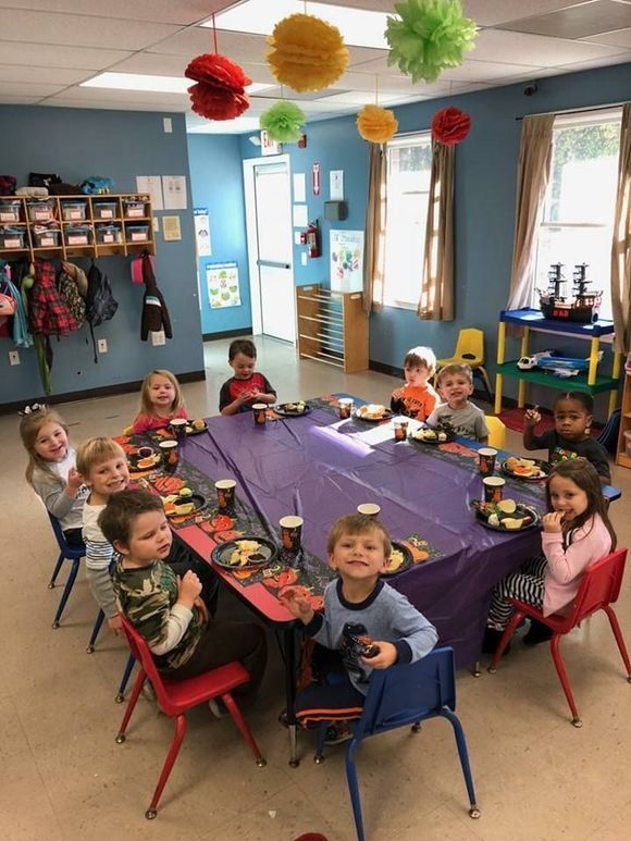 Children seated around a purple table in a classroom, with colorful hanging decorations overhead.