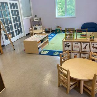 Preschool classroom with small tables, play kitchen, alphabet rug, and animal blocks by a window