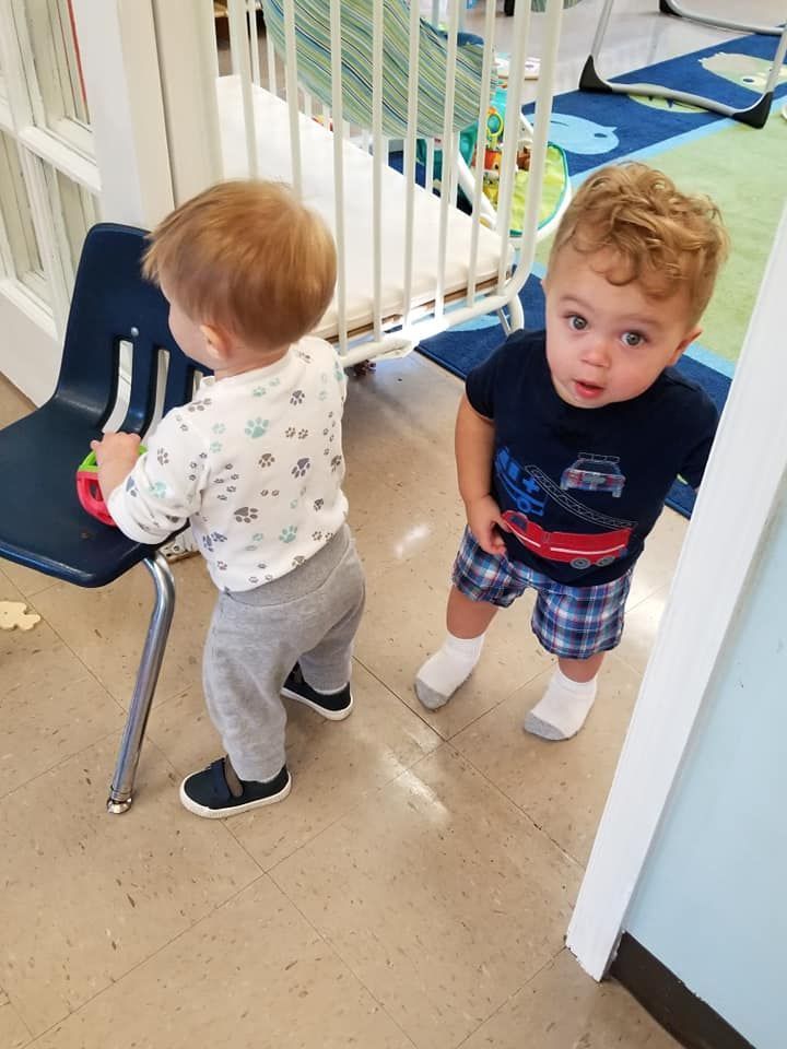 Two toddlers in a play area, one standing by a chair and one looking up near a doorway.