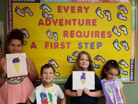 Three children holding art projects in front of a yellow poster reading “Every adventure requires a first step”