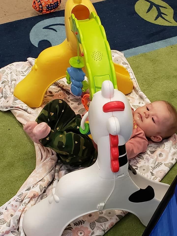 Baby lying on a blanket beside colorful plastic toy slides and an animal rocker in a play area.