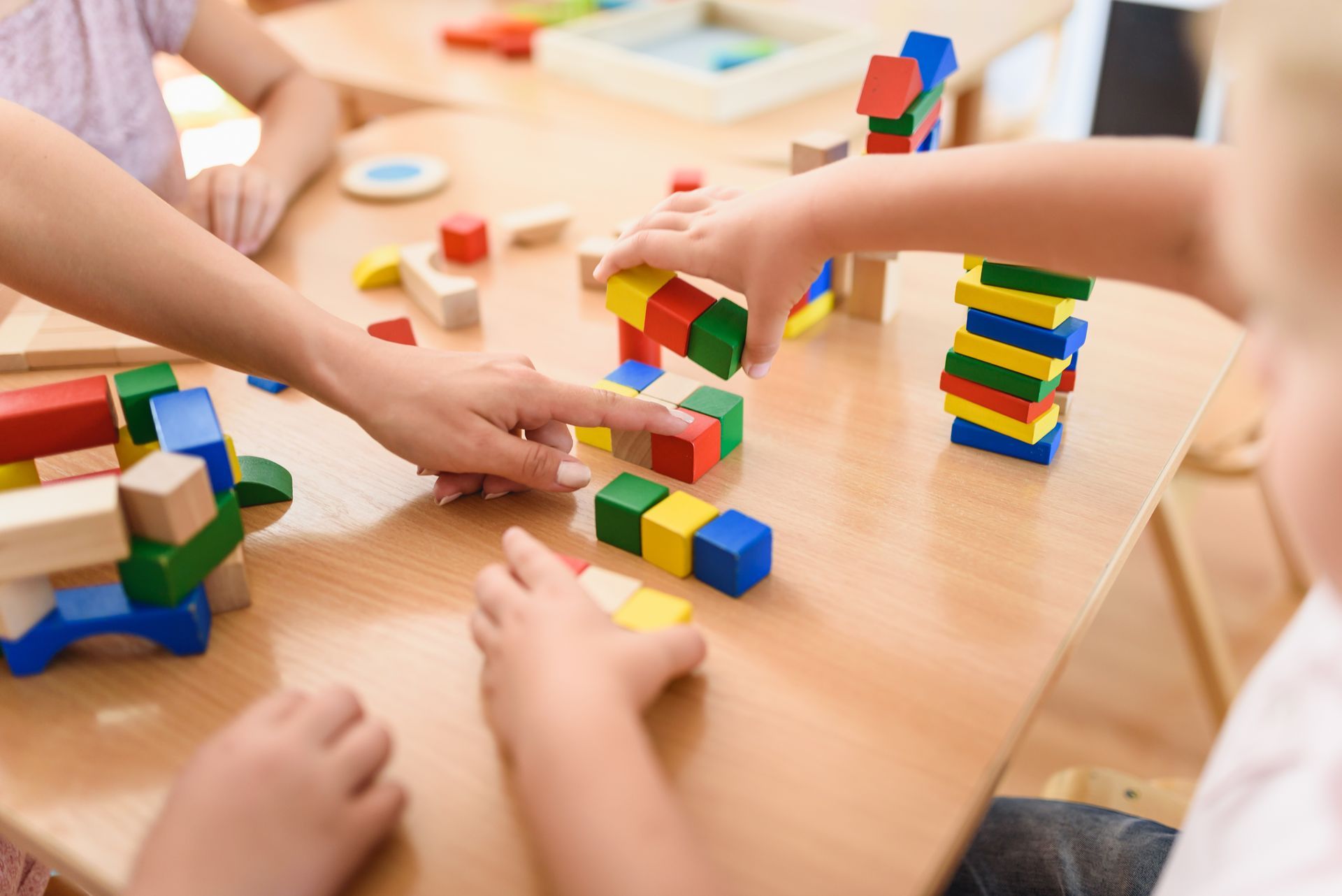 Children building colorful block towers on a table, with hands reaching in to play together.
