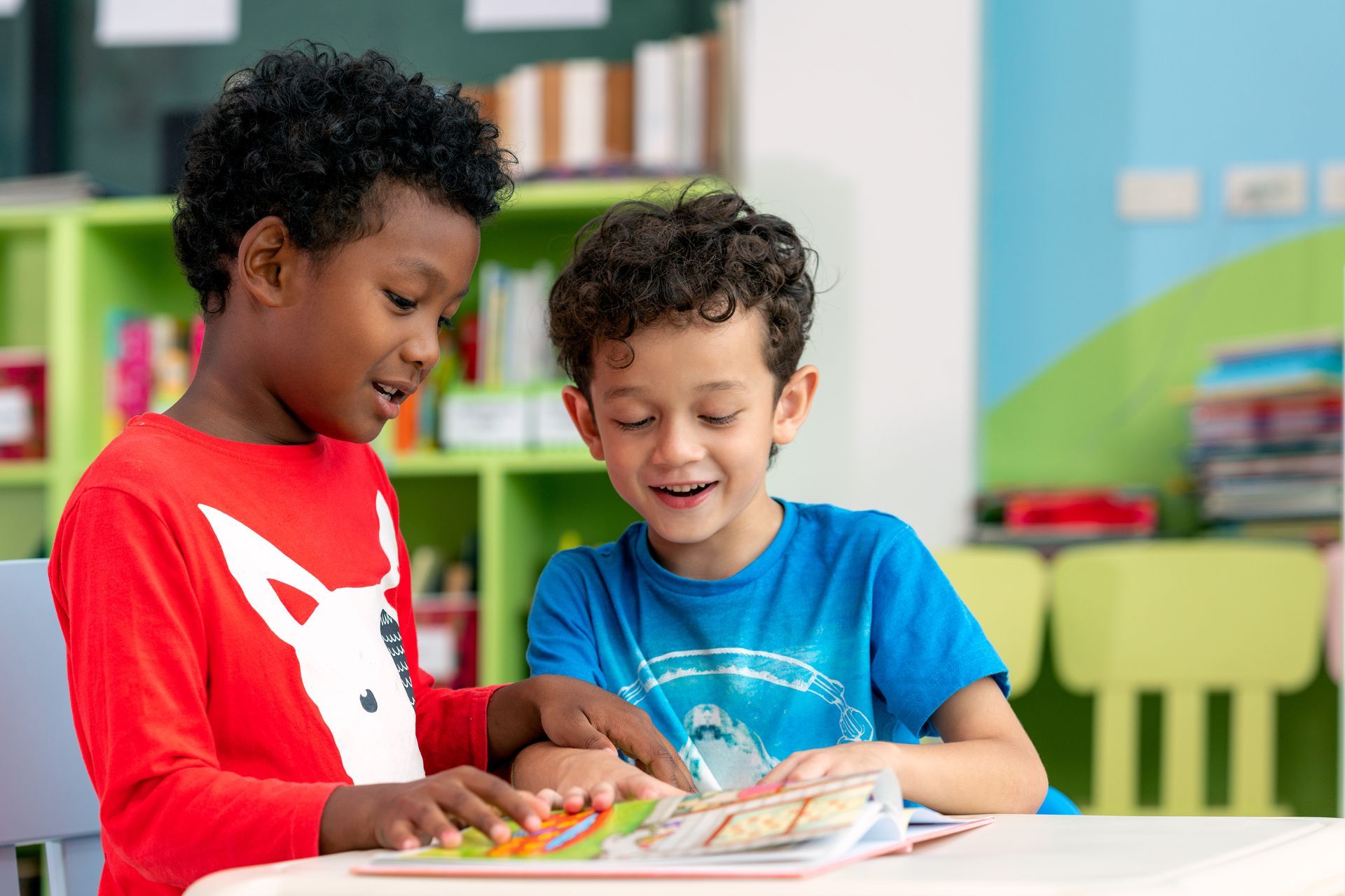 Two children smiling and reading a book together at a table in a colorful classroom