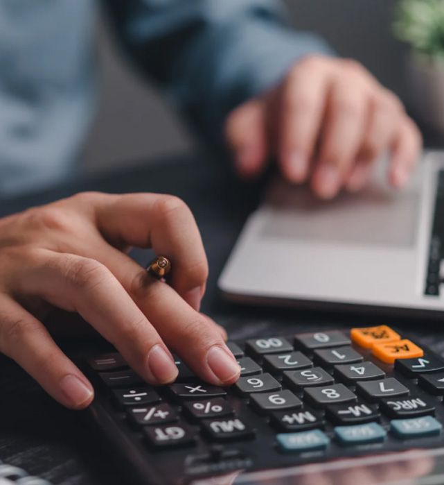 Hands using a calculator near a laptop keyboard. Fingers press buttons, orange