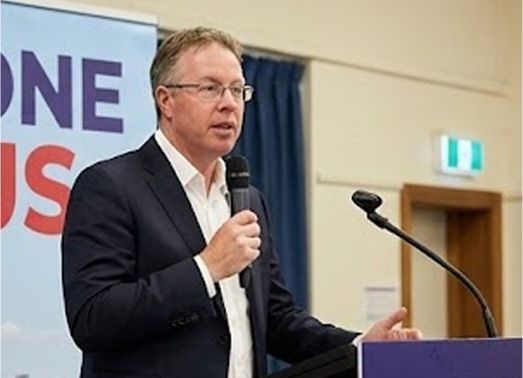 Man in a suit speaks into a microphone at a podium. A banner with text is in the background.
