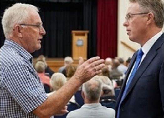Two men in conversation at a public meeting. One gestures, the other listens. Audience in the background.