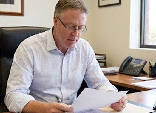 Man in glasses examines papers at a desk in an office setting.