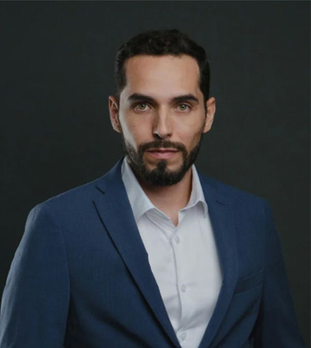 Man in blue suit, white shirt, looking forward, serious expression, dark background.
