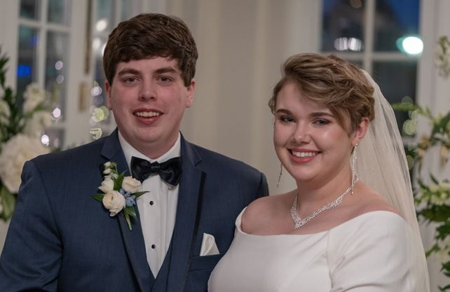 Couple in wedding attire smiling, indoors; the man wears a navy suit, the woman a white gown and veil.