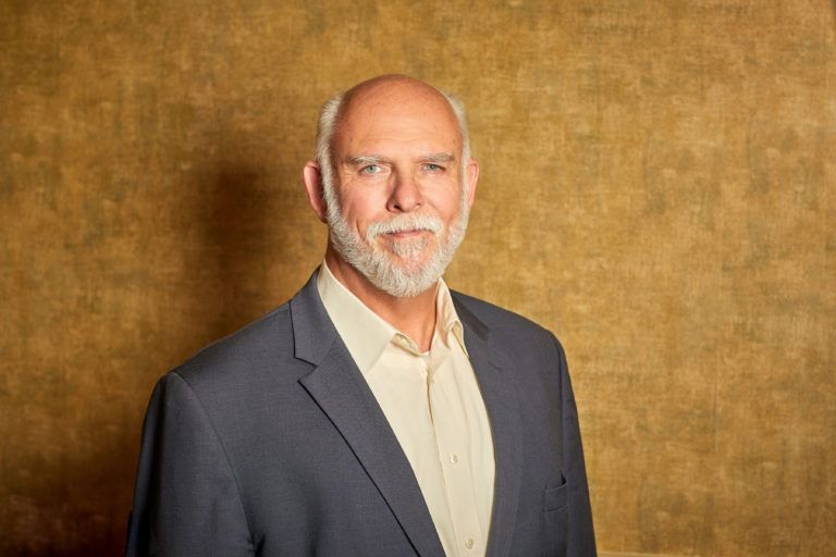 A man in a suit and white shirt is standing in front of a brown wall.