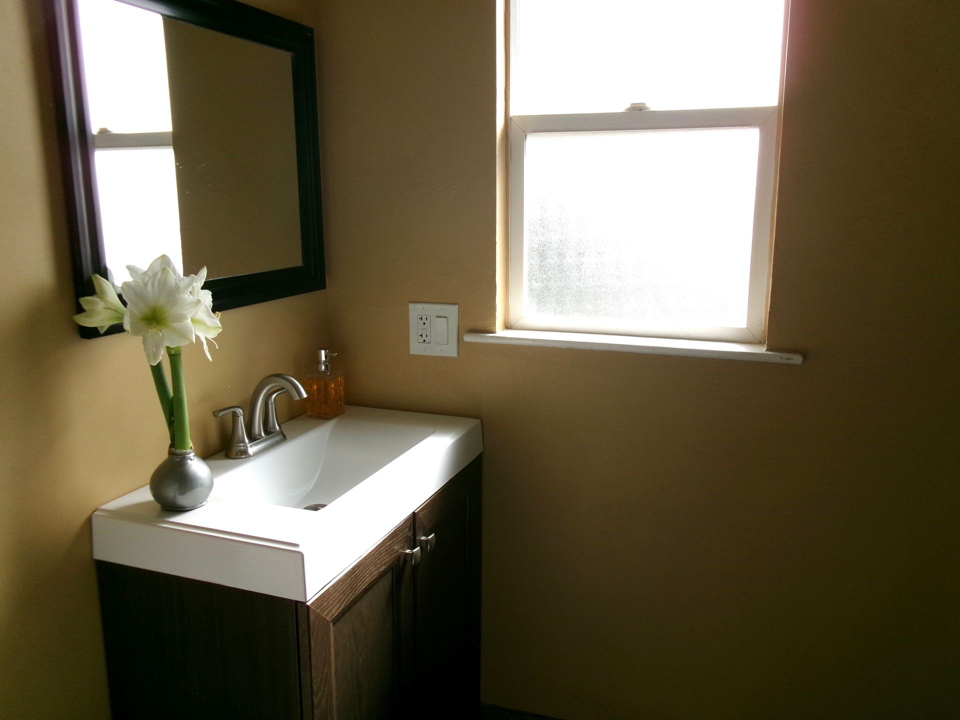 A dark wood vanity with a white basin, a mirror, and a vase of white flowers next to a window in a tan bathroom.