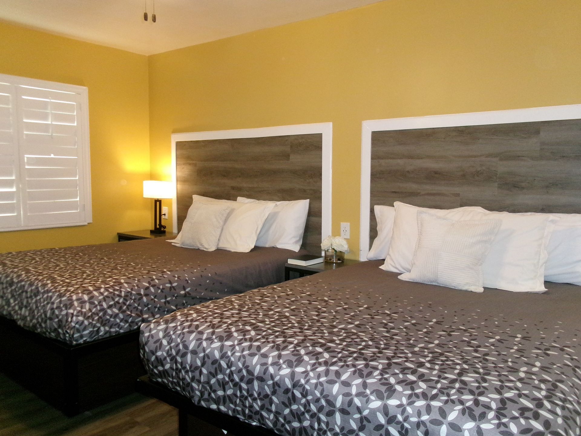A hotel room with two beds, featuring gray patterned quilts, wood-look headboards, and yellow walls.