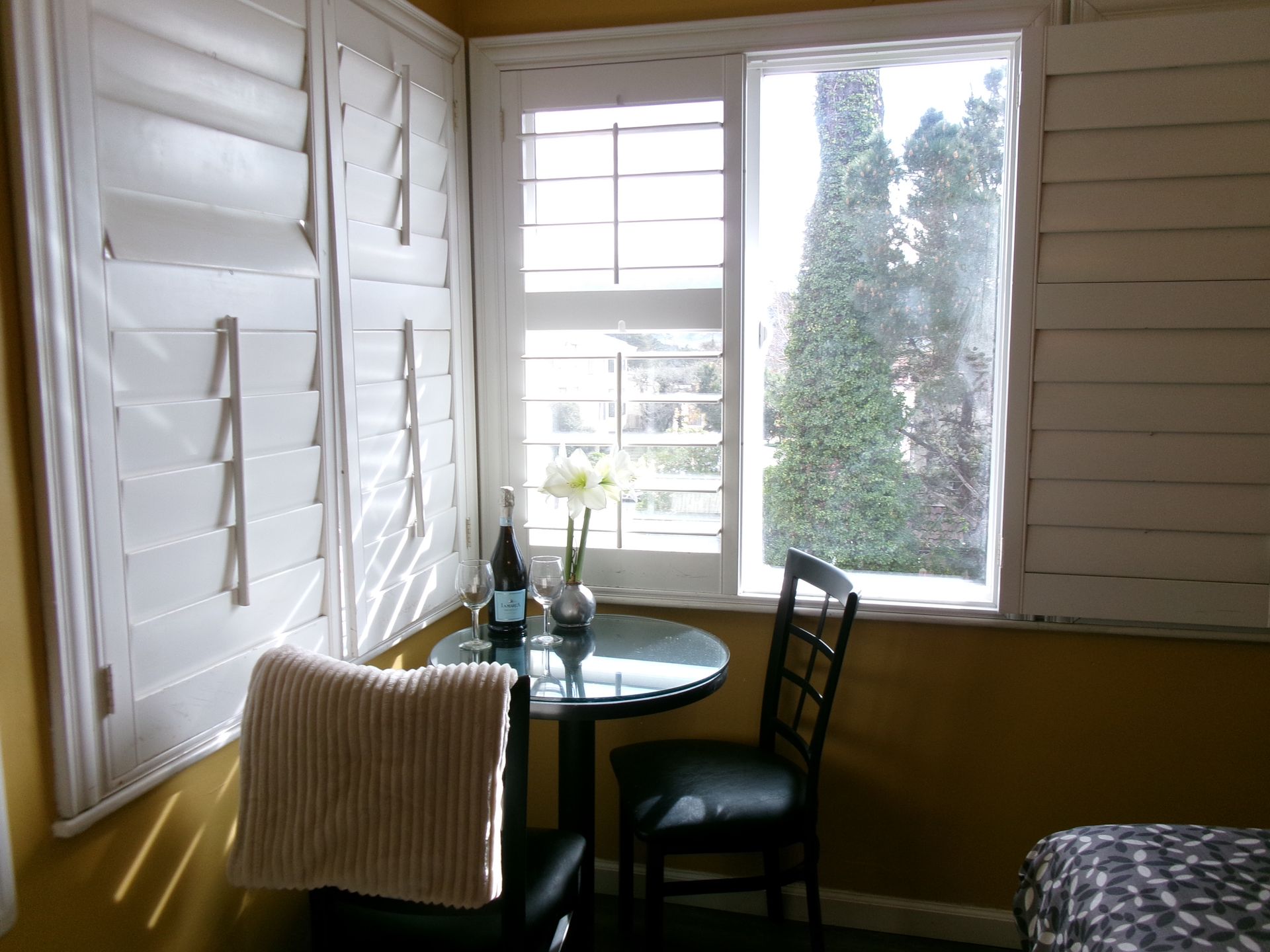 A small round glass table with a vase of flowers and a bottle, set by a window with white shutters in a yellow room.