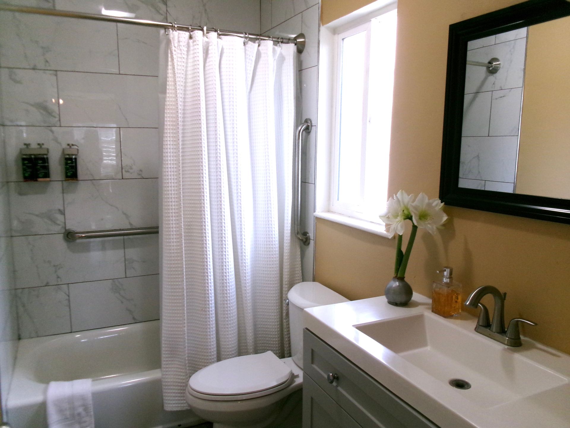 A bright bathroom featuring a white marble-tiled bathtub with a shower, a grey vanity with a white sink, and a mirror.