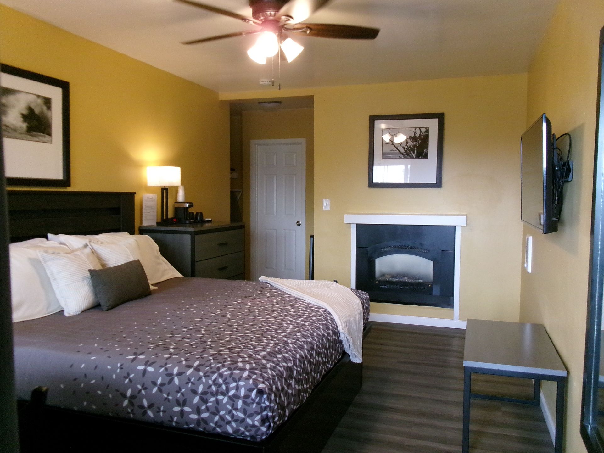 A hotel room with a queen bed featuring a patterned quilt, a fireplace, television, desk, and mustard yellow walls.