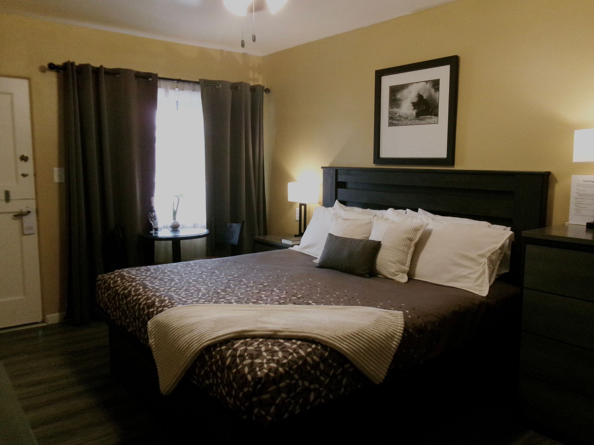 A hotel room with a queen bed featuring a dark patterned comforter, dark curtains, and a framed wall art above the head.