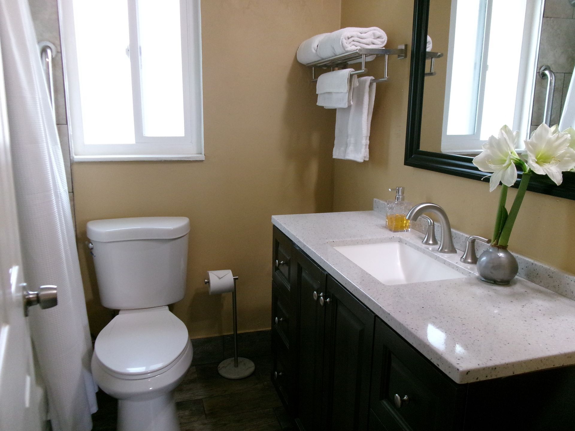 A bathroom with a white toilet, dark vanity with a speckled countertop, a mirror, and a small vase of white flowers.