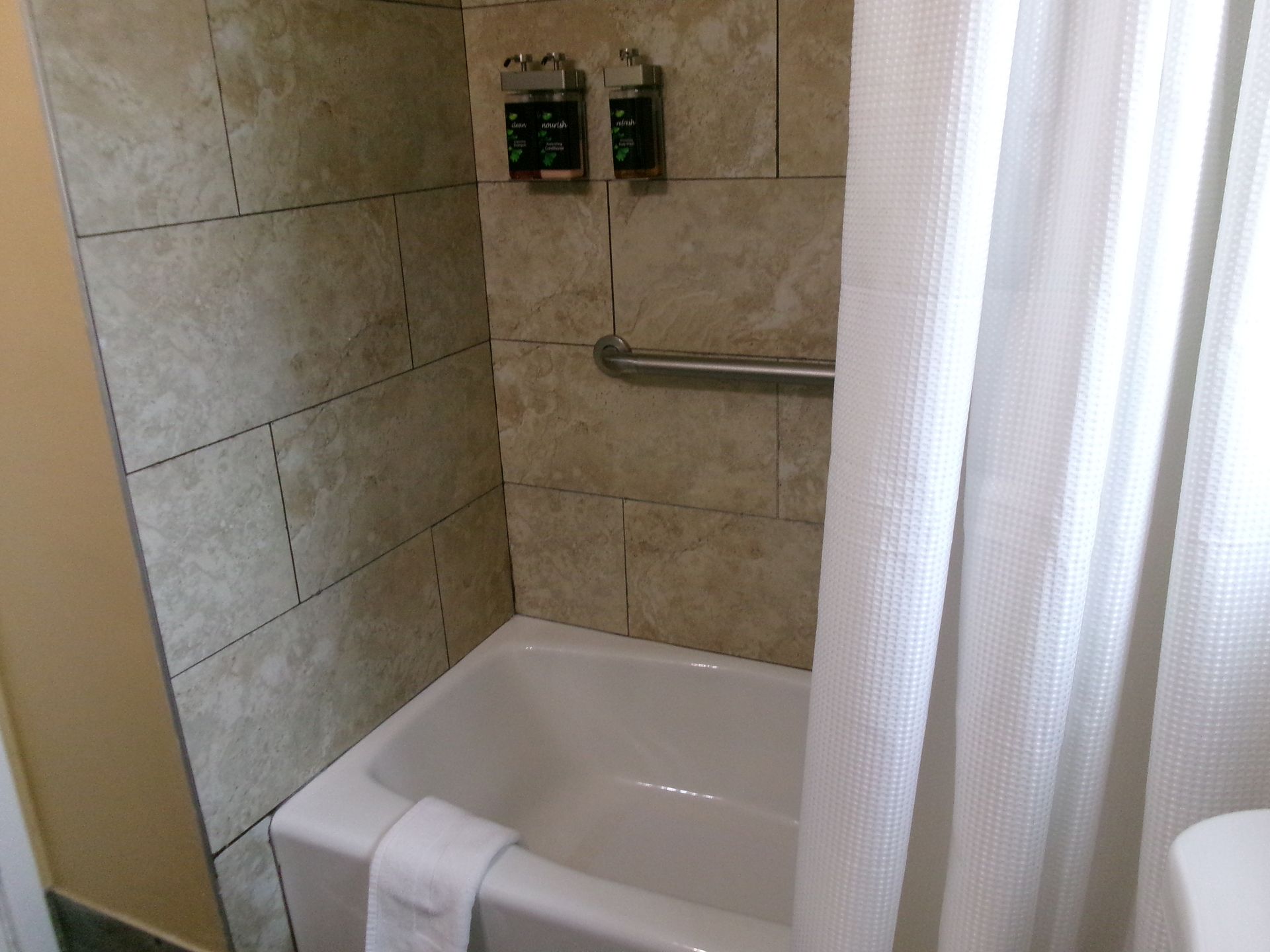 A hotel bathtub with light-colored tile walls, a grab bar, two wall-mounted dispensers, and a white shower curtain.