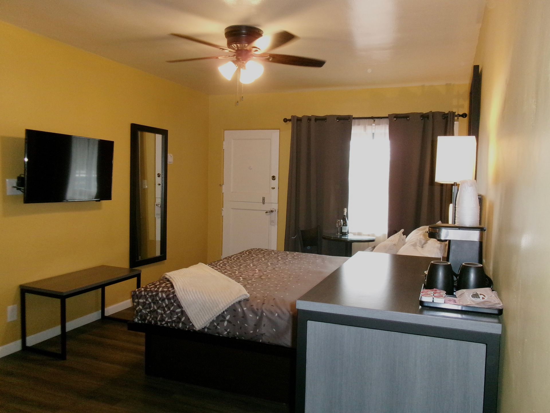 A cozy, yellow-walled hotel room featuring a queen bed, flat-screen TV, ceiling fan, and a kitchenette area.