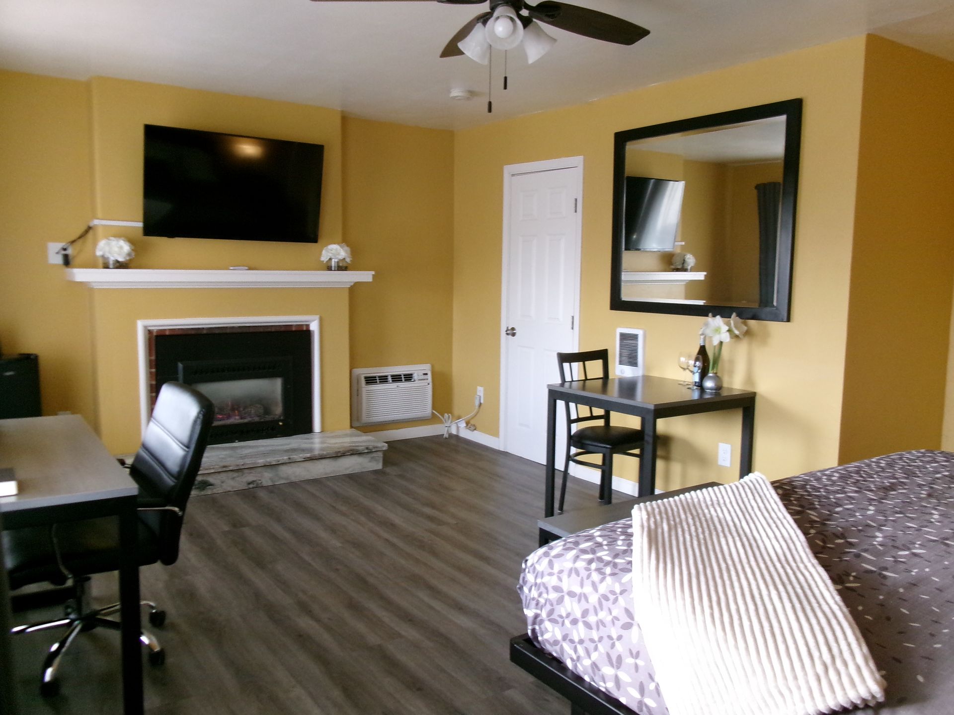 A bedroom with yellow walls, a fireplace, television, desk, and dining table, featuring dark flooring and a ceiling fan.
