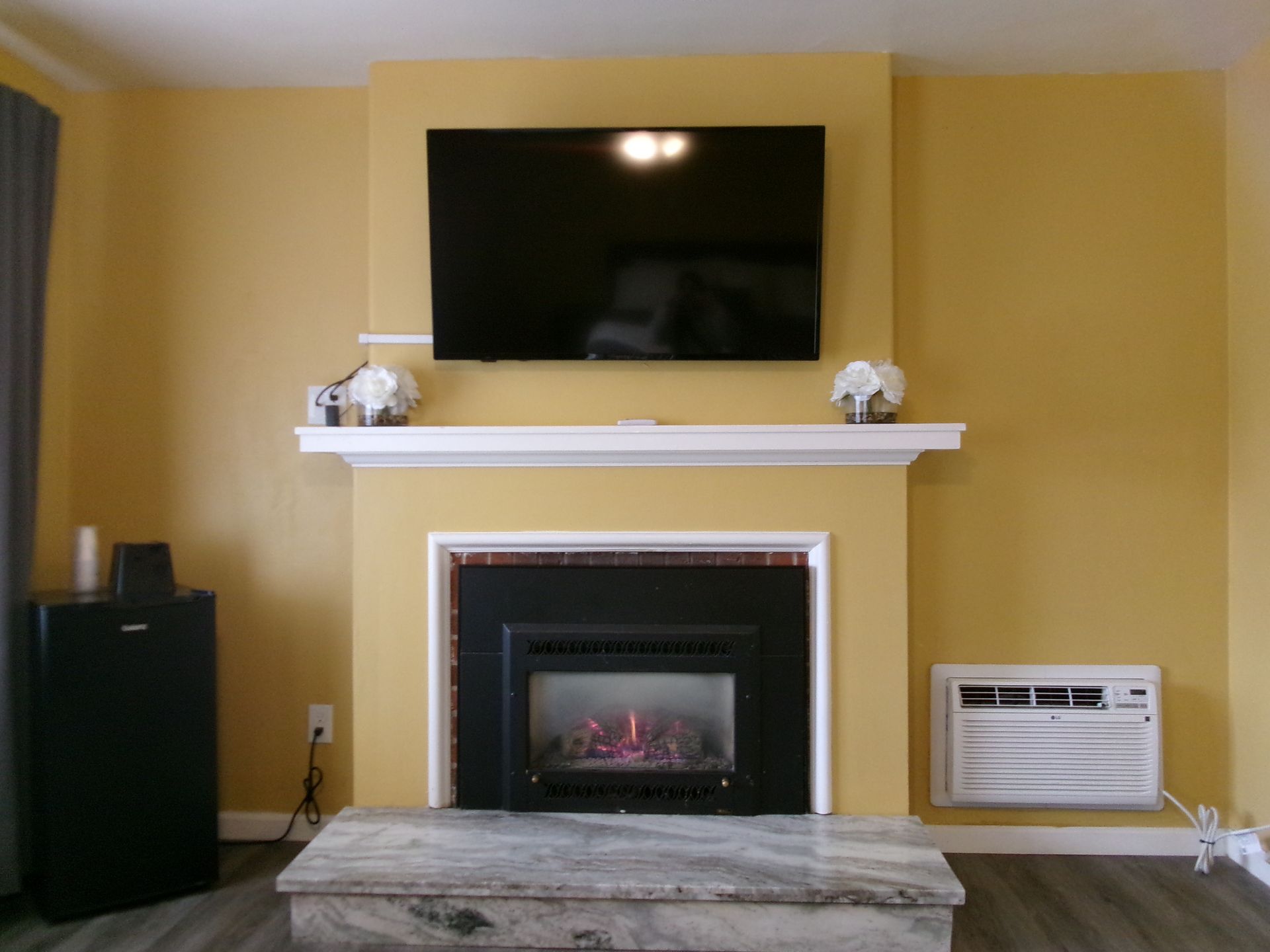A wall-mounted TV above a white fireplace mantel, with a marble hearth and a wall air conditioning unit to the right.
