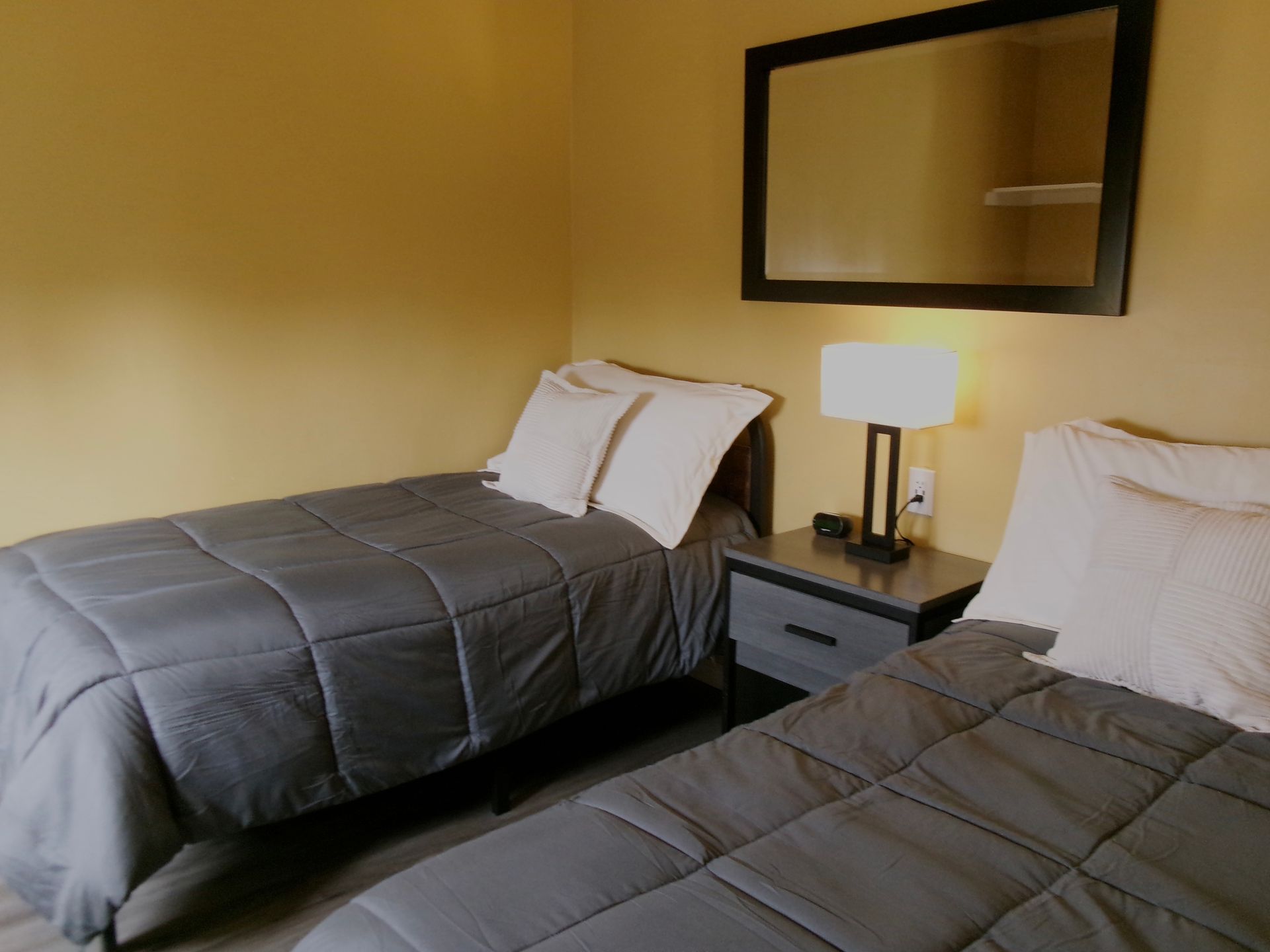 Two twin beds with gray comforters and white pillows, separated by a nightstand with a lamp and a mirror on the wall.