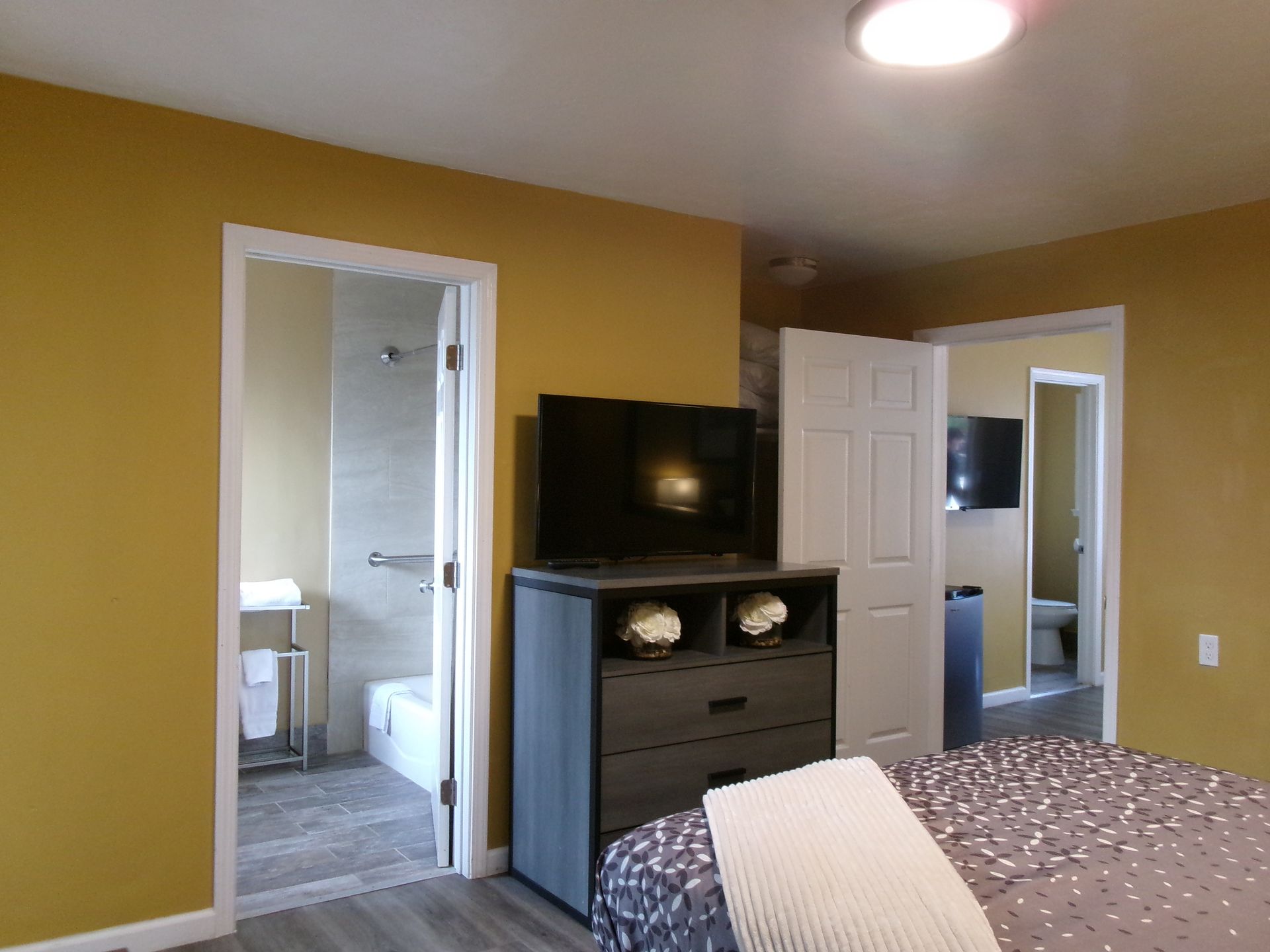 A bedroom with mustard-yellow walls, a gray dresser topped with a TV, a bed, and doorways leading to two bathrooms.