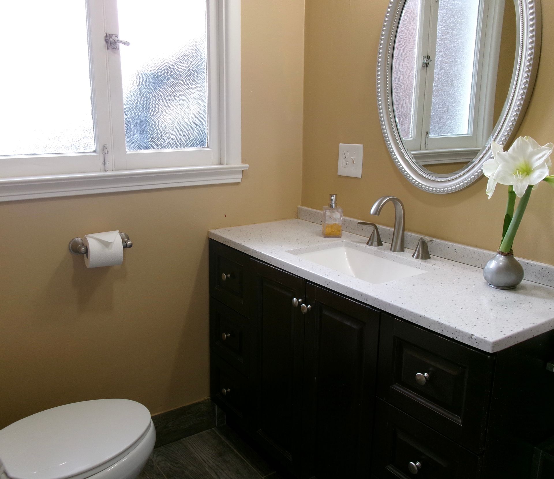 A bathroom vanity with a dark wood cabinet, white quartz countertop, oval mirror, and a flower vase, next to a toilet.