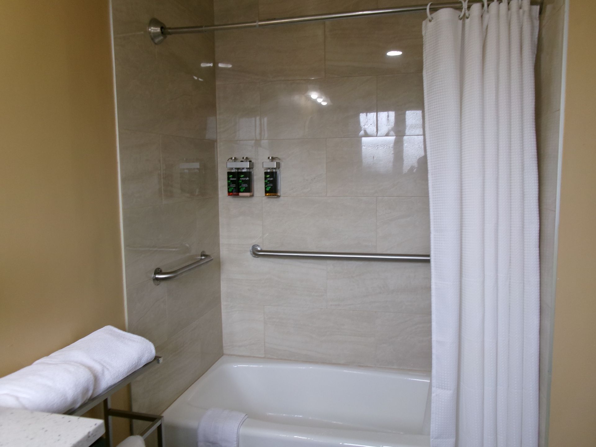 A beige-tiled hotel shower area with a bathtub, white curtain, grab bars, and wall-mounted dispensers.