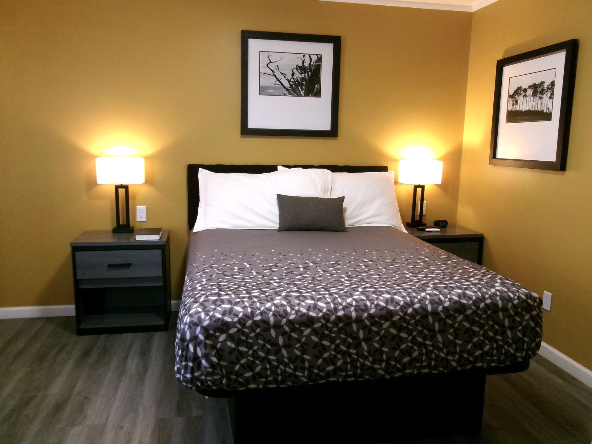 A bedroom with a bed featuring a patterned gray quilt, two nightstands with lamps, and framed art on mustard-yellow walls.