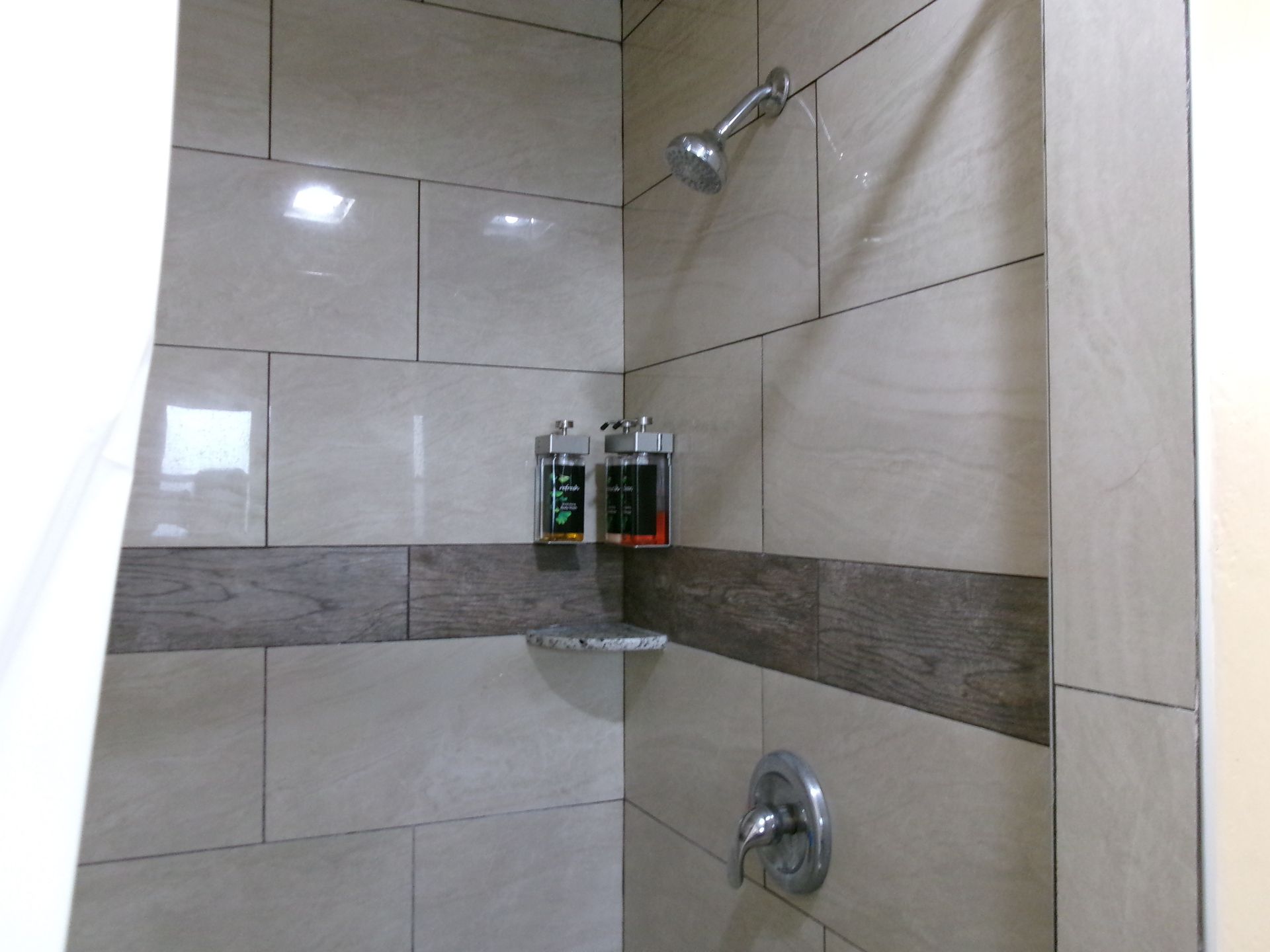 Shower stall with light-colored tiles, a wood-look accent border, a chrome shower head, faucet, and wall-mounted dispensers.