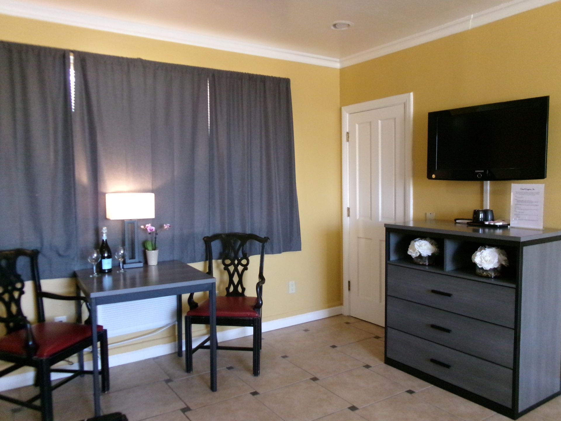 A small dining area with a table, two chairs, a lamp, and a dresser topped with a TV in a room with yellow walls.