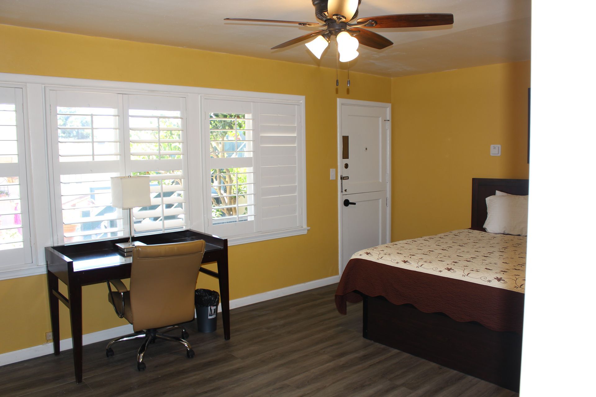 A bedroom with yellow walls, a brown bed frame, a white bedspread, a wooden desk with a tan chair, and white shutters.