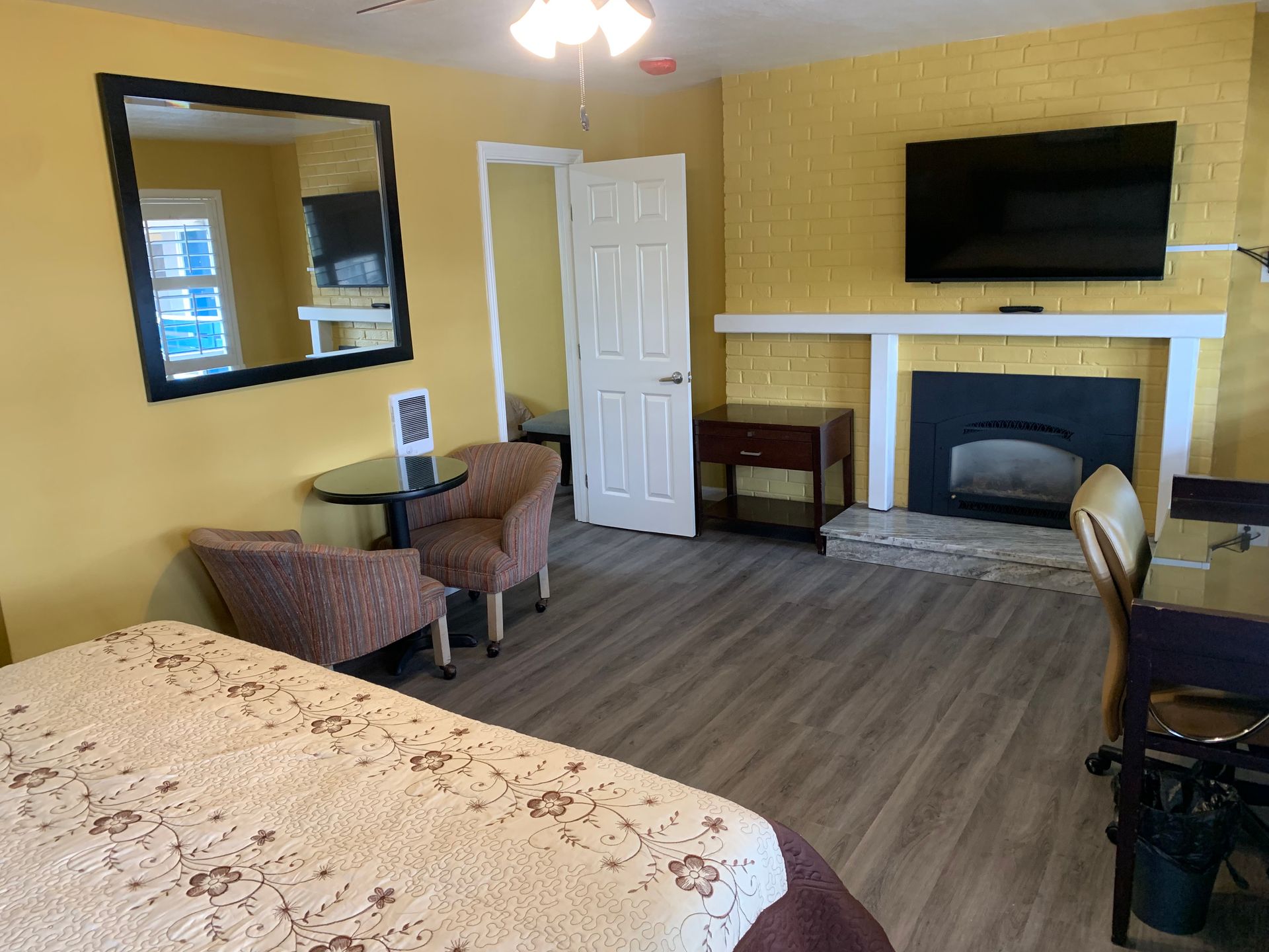 A hotel room with yellow walls, a king bed, a fireplace with a mounted television, a small table, chairs, and a mirror.