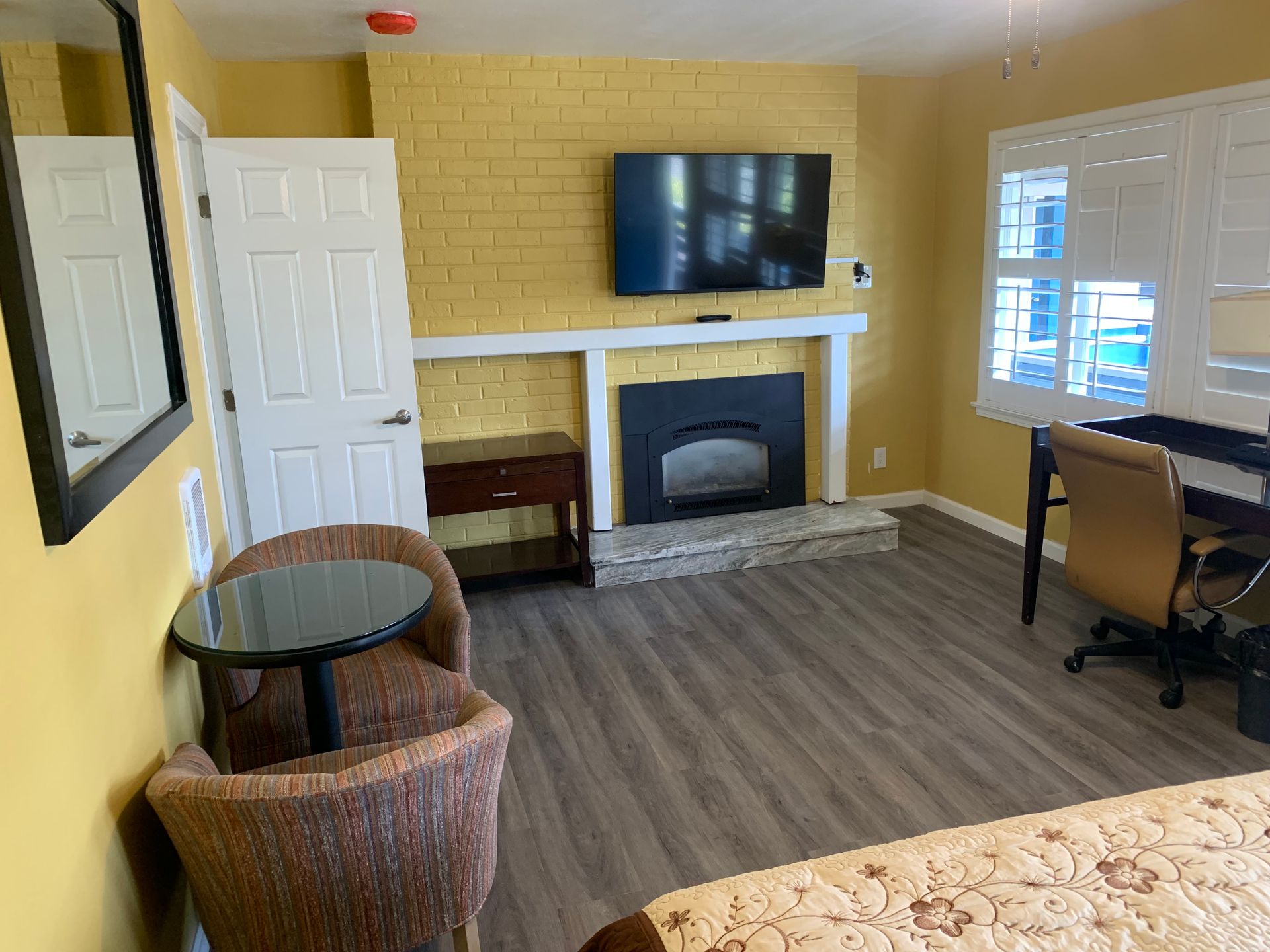 A hotel room with yellow walls, a fireplace with a mounted television, a small round table with two chairs, and a desk.