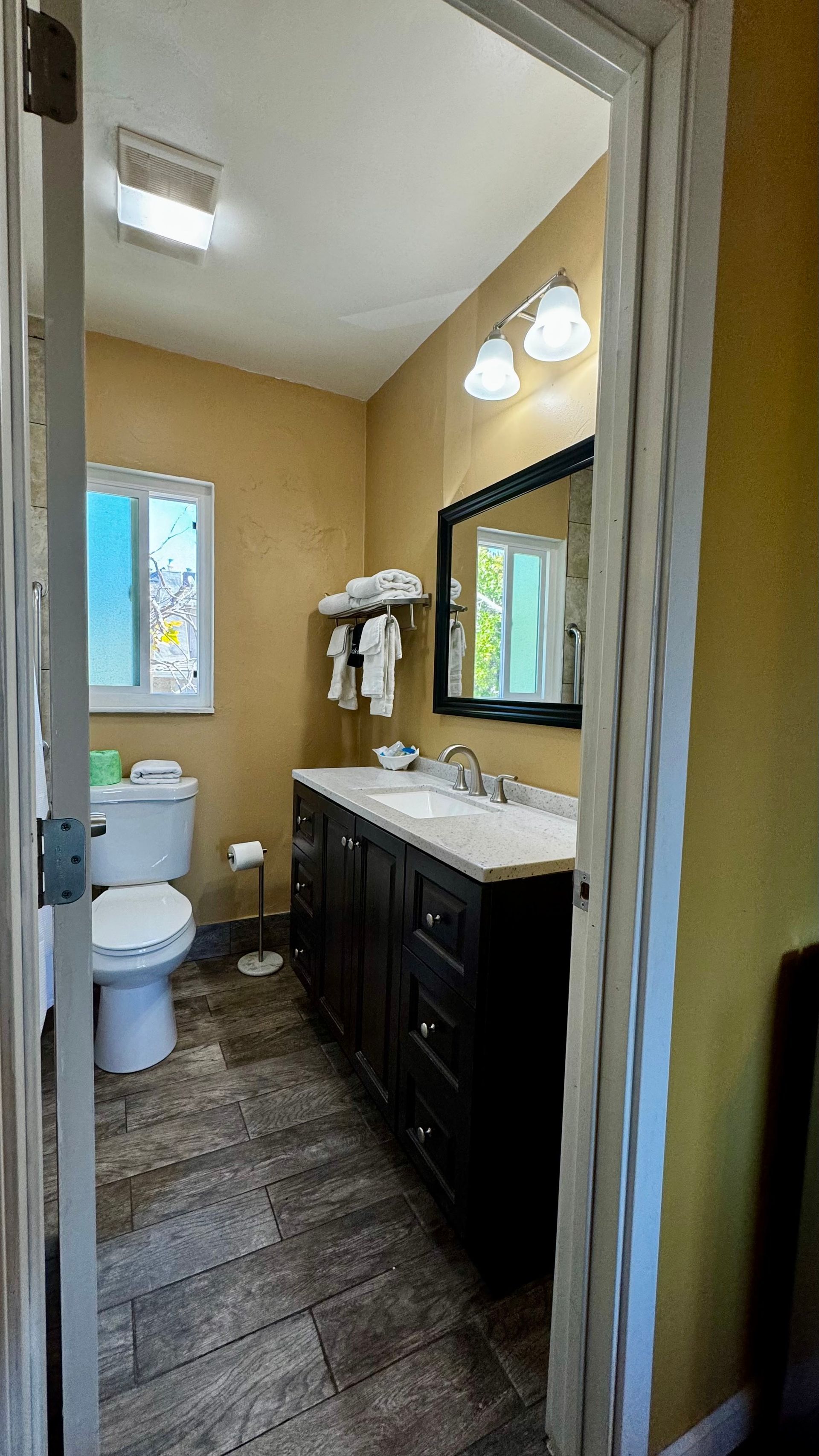 A bathroom with tan walls, dark cabinetry, a white toilet, stone-tiled floors, and a vanity mirror with lights above.