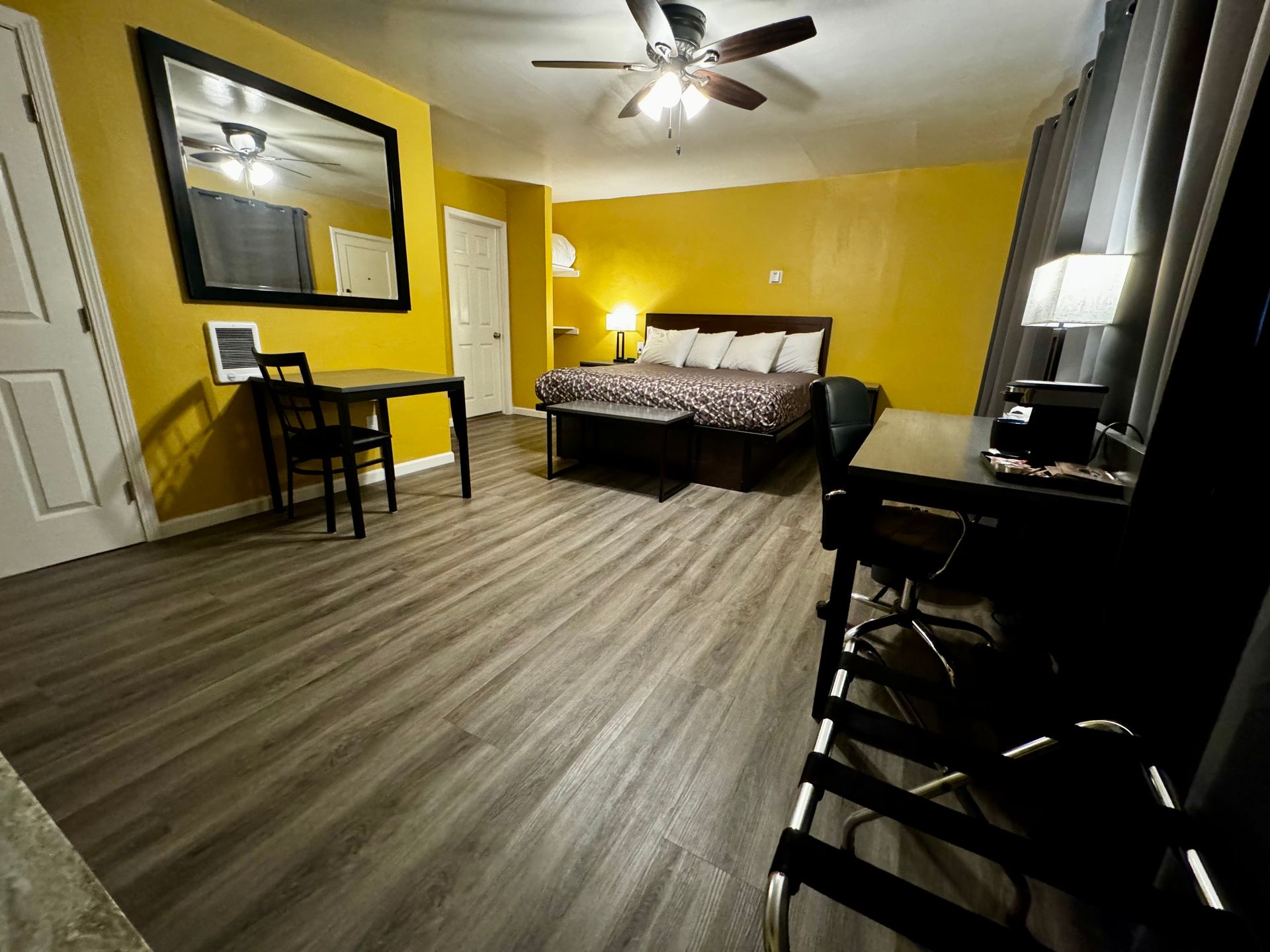 A hotel room with wood floors, yellow walls, a bed, a desk, a dining set, and a large mirror.
