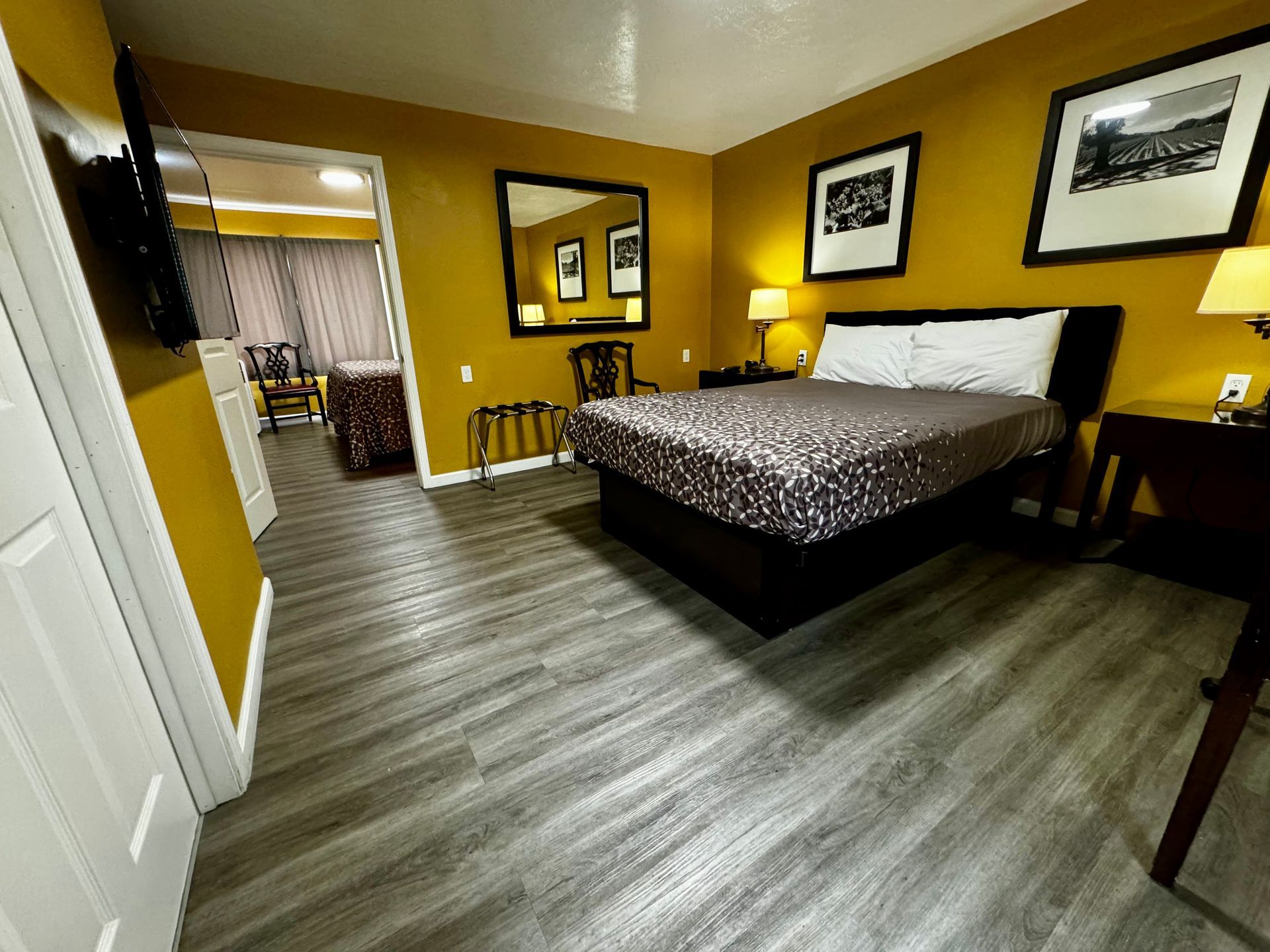 A hotel room with yellow walls, a queen-sized bed, grey wood-look flooring, two framed prints, and an adjacent room view.