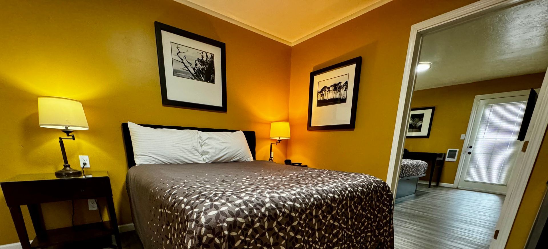 A bedroom with a patterned bedspread, bedside lamp, two framed artworks on yellow walls, and a doorway to a second room.