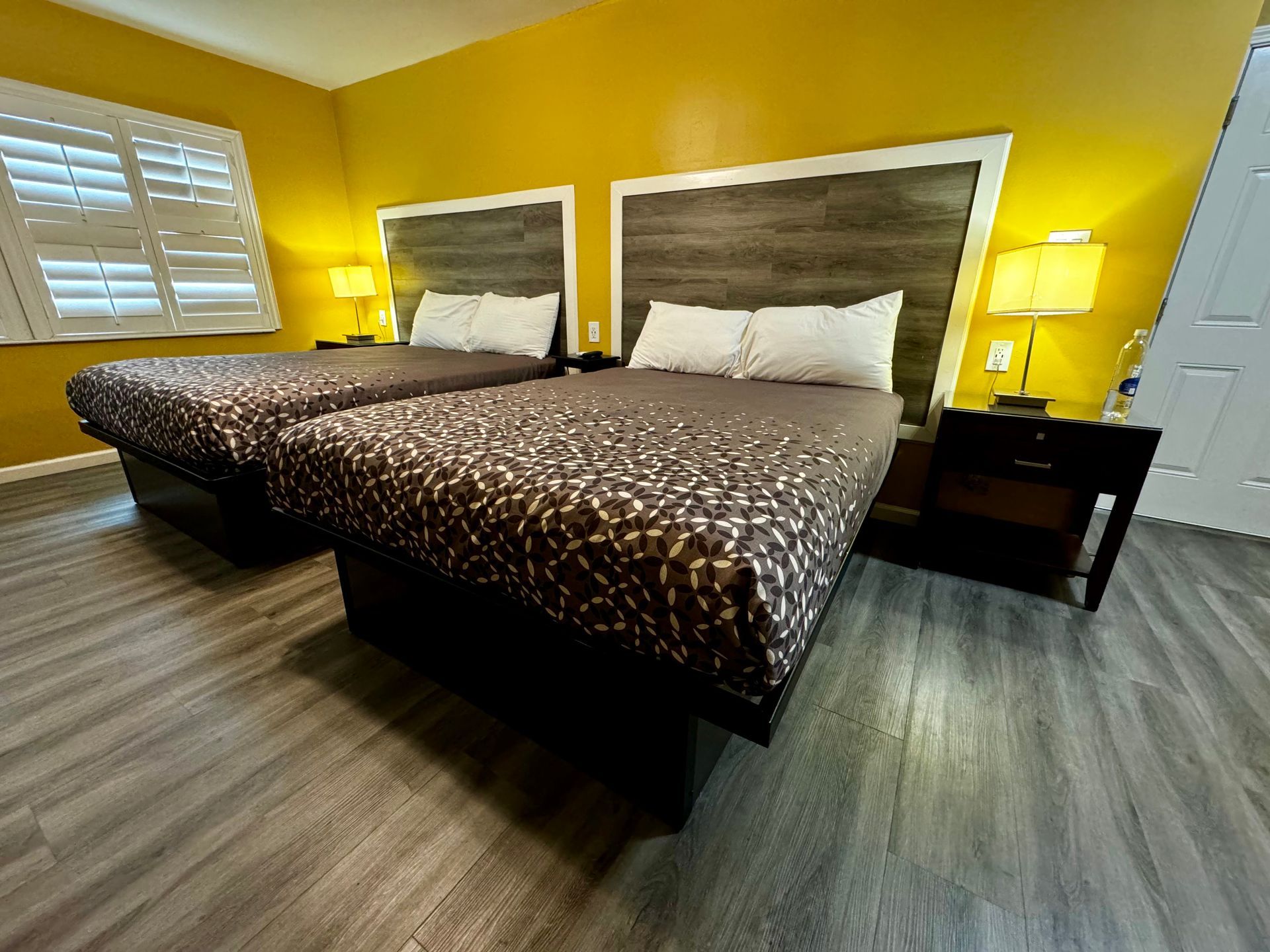 A hotel room with two queen beds, dark wood headboards, yellow walls, and light grey wood-look flooring.