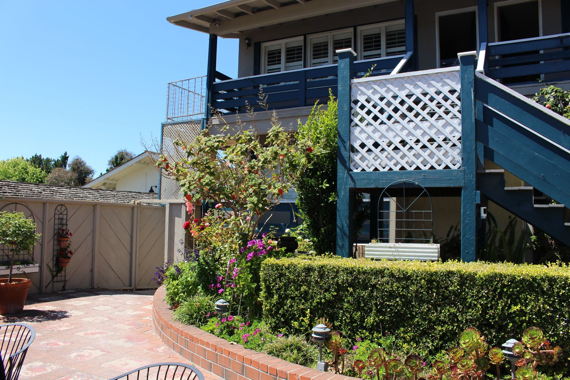 A two-story building with a dark blue wooden staircase and lattice railing, alongside a garden and brick-paved patio.