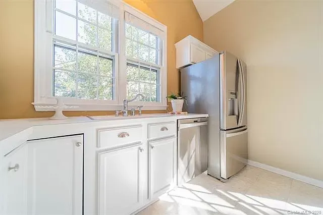 A kitchen with white cabinets , a stainless steel refrigerator, a sink, and a window.