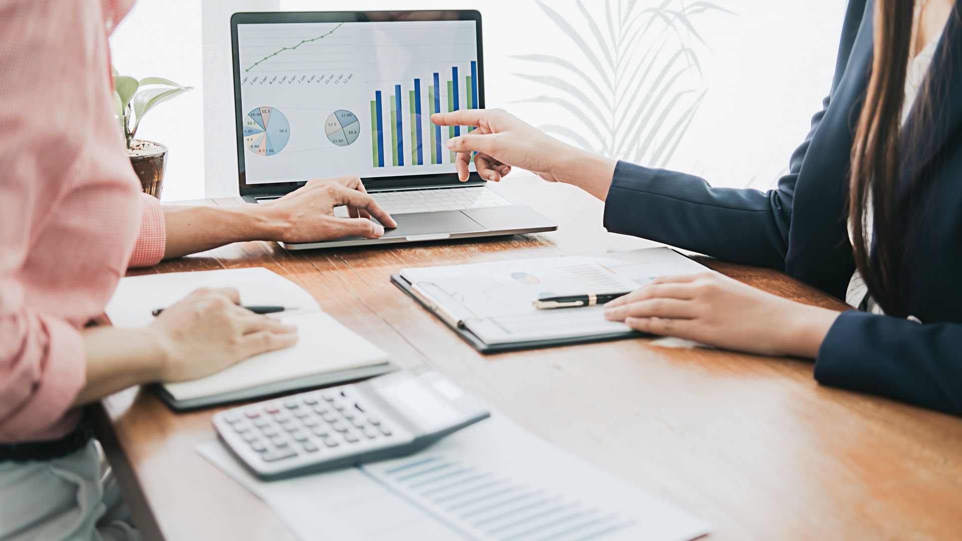 Two people review financial data on a laptop, pointing at charts, with a calculator and notepad on a desk. Two people review financial data on a laptop, pointing at charts, with a calculator and notepad on a desk.