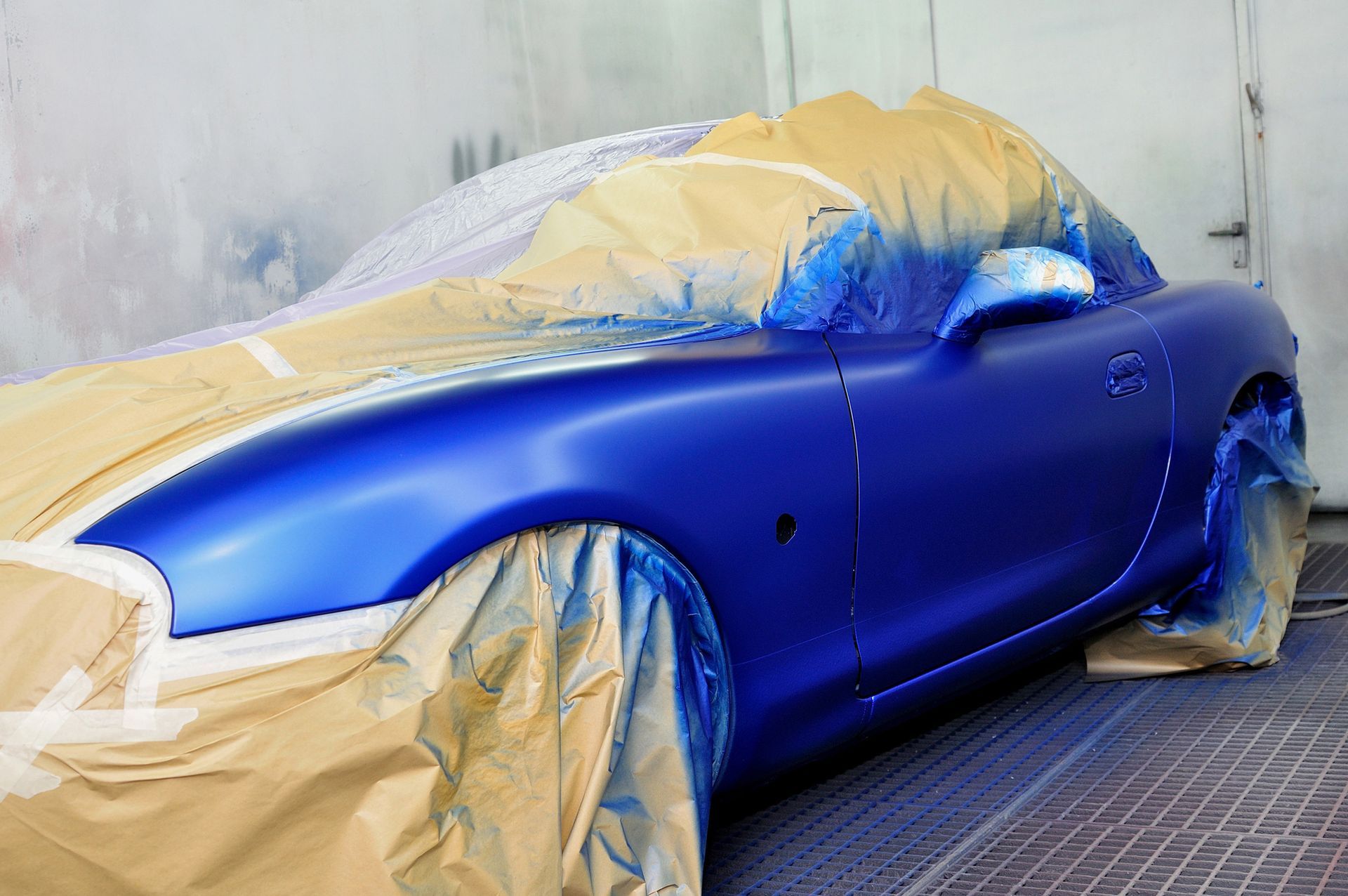 Un'auto sportiva blu brillante ricoperta di carta protettiva durante una verniciatura professionale.